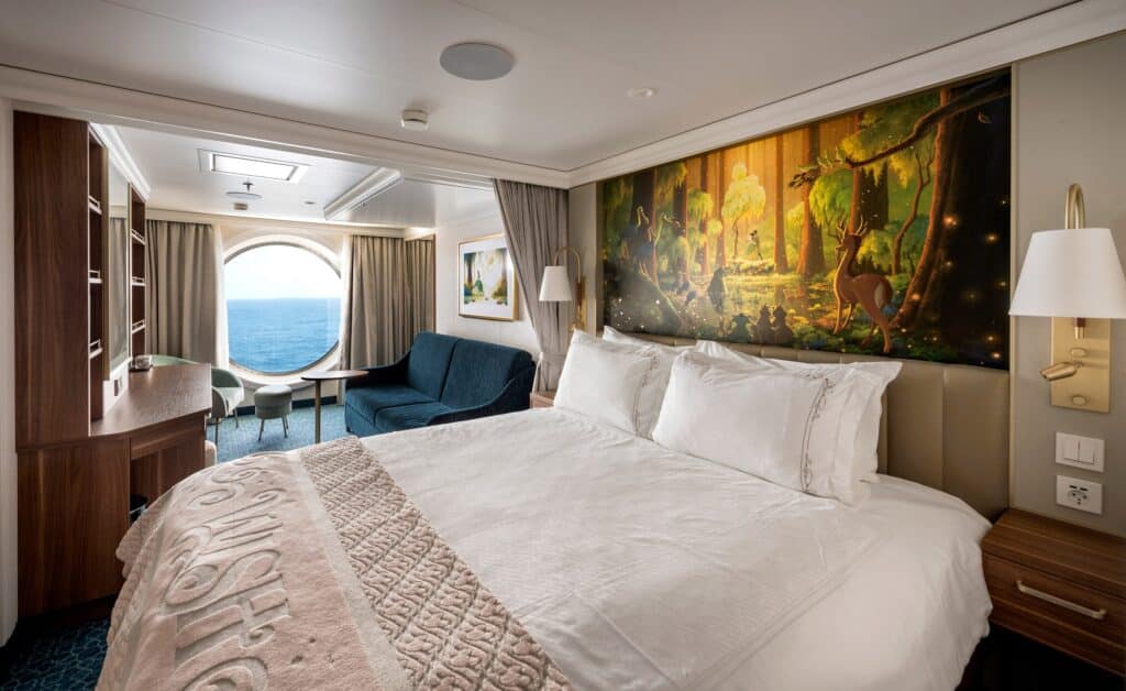 Deluxe oceanview stateroom del Disney Wish. Una cama queen con edredón blanco, una pintura de un busque en la cabecera y al fondo una ventana redonda con vista al mar. Es un ejemplo de uno de los tipos de camarote de los cruceros de Disney