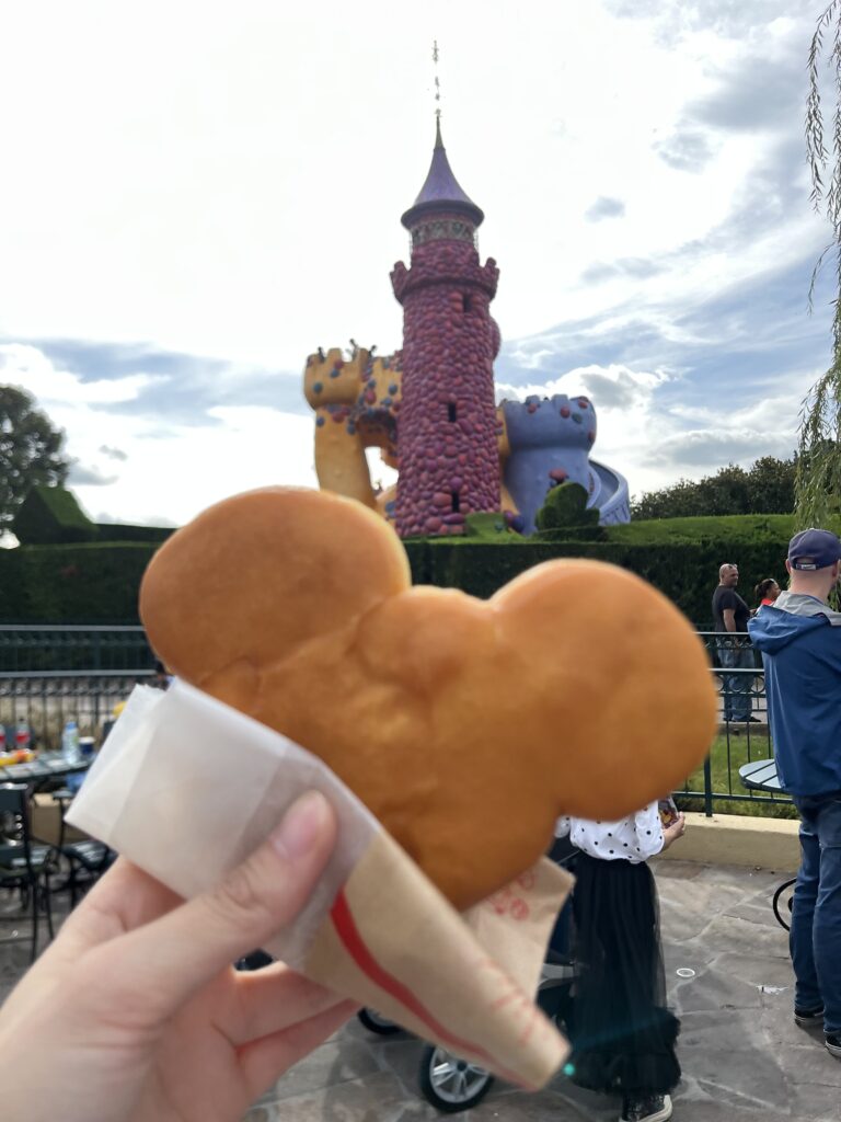 Beignet con forma de Mickey Mouse con el castillo del laberinto de Alicia en el País de las Maravillas de fondo. Snack que puedes encontrar en The Old Mill, uno de los mejores restaurantes de Disneyland Paris