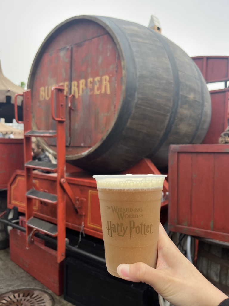 Cerveza de mantequilla y de fondo el puesto de butterbeer en el Mundo de Harry Potter en Universal Orlando